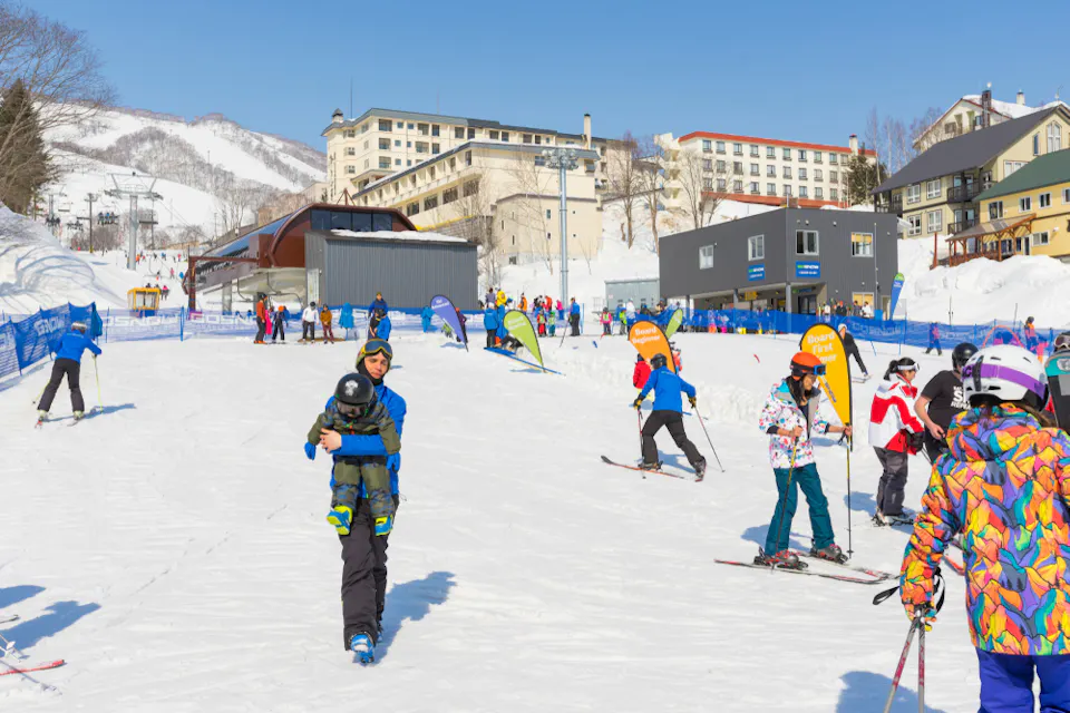 A snowy ski resort with people skiing and snowboarding. A person in the foreground carries a child. The background features ski lifts, buildings, and mountains. The scene is lively with various people enjoying winter activities.