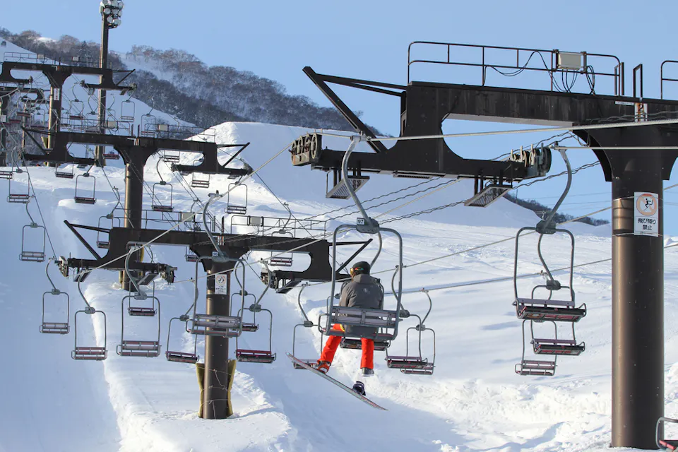 A skier in red pants sits on a ski lift ascending a snowy mountain under a clear blue sky. The image shows multiple empty ski lift chairs, snow-covered slopes, and leafless trees in the background.