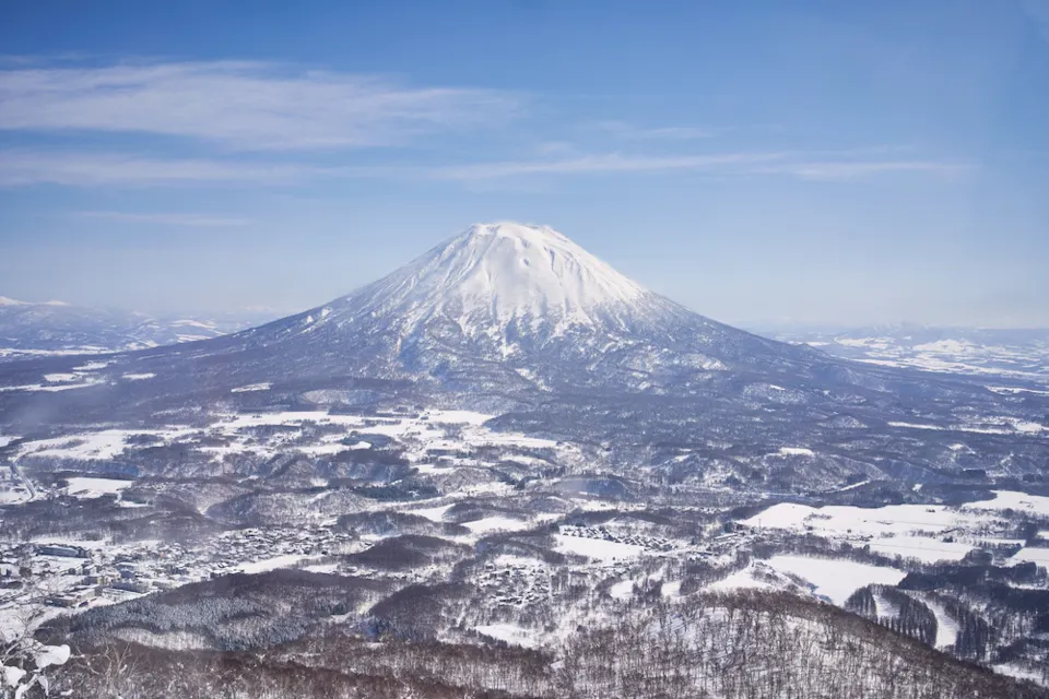 Snow-covered Mount Yōtei in Hokkaido, Japan, stands tall under a clear blue sky. The surrounding landscape is blanketed in snow, with scattered trees and small buildings visible in the foreground.