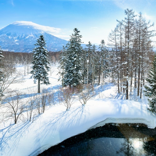 Grand Hirafu Onsens A snow-covered landscape with tall evergreen trees under a clear blue sky. A distant mountain rises in the background. In the foreground, a calm, partially frozen body of water reflects the surrounding scenery.