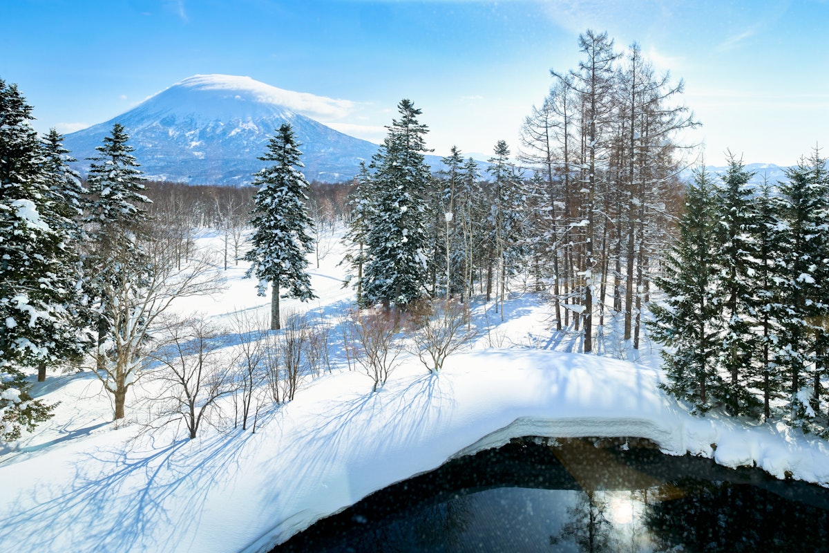 Grand Hirafu Onsens A snow-covered landscape with tall evergreen trees under a clear blue sky. A distant mountain rises in the background. In the foreground, a calm, partially frozen body of water reflects the surrounding scenery.