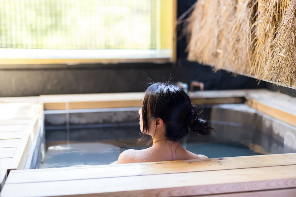 A person with dark hair sits in a wooden hot tub, partially submerged in water, with a straw-covered wall to the right and a window in the background letting in natural light.