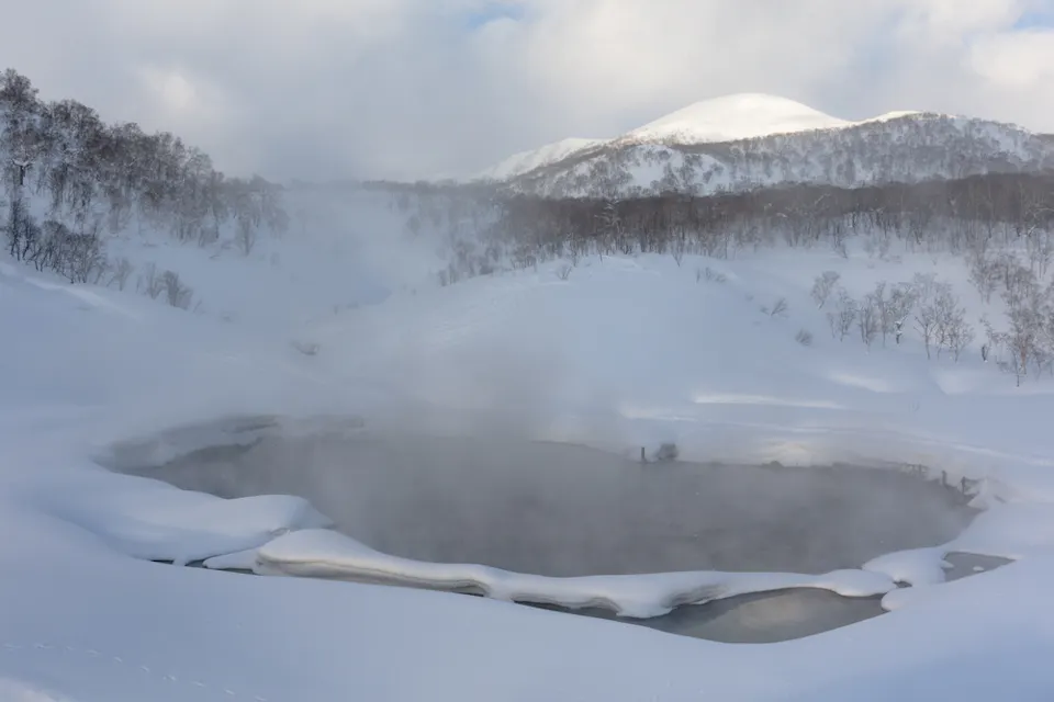 Snow-covered landscape with a steaming hot spring in the foreground. Trees are dusted with snow, and a mountain looms in the background under a cloudy sky. The scene is serene and predominantly white and gray.