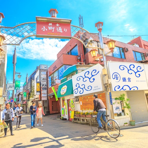 Kamakura Street A vibrant street scene in a Japanese town, featuring a cyclist and pedestrians. The area is lined with colorful shops and signs under a decorative archway, with a bright blue sky overhead.