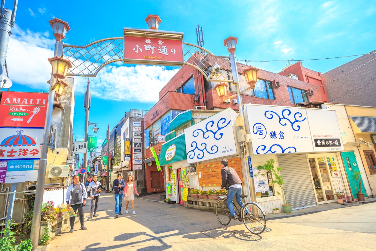 Kamakura Street A vibrant street scene in a Japanese town, featuring a cyclist and pedestrians. The area is lined with colorful shops and signs under a decorative archway, with a bright blue sky overhead.