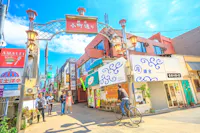A vibrant street scene in a Japanese town, featuring a cyclist and pedestrians. The area is lined with colorful shops and signs under a decorative archway, with a bright blue sky overhead.