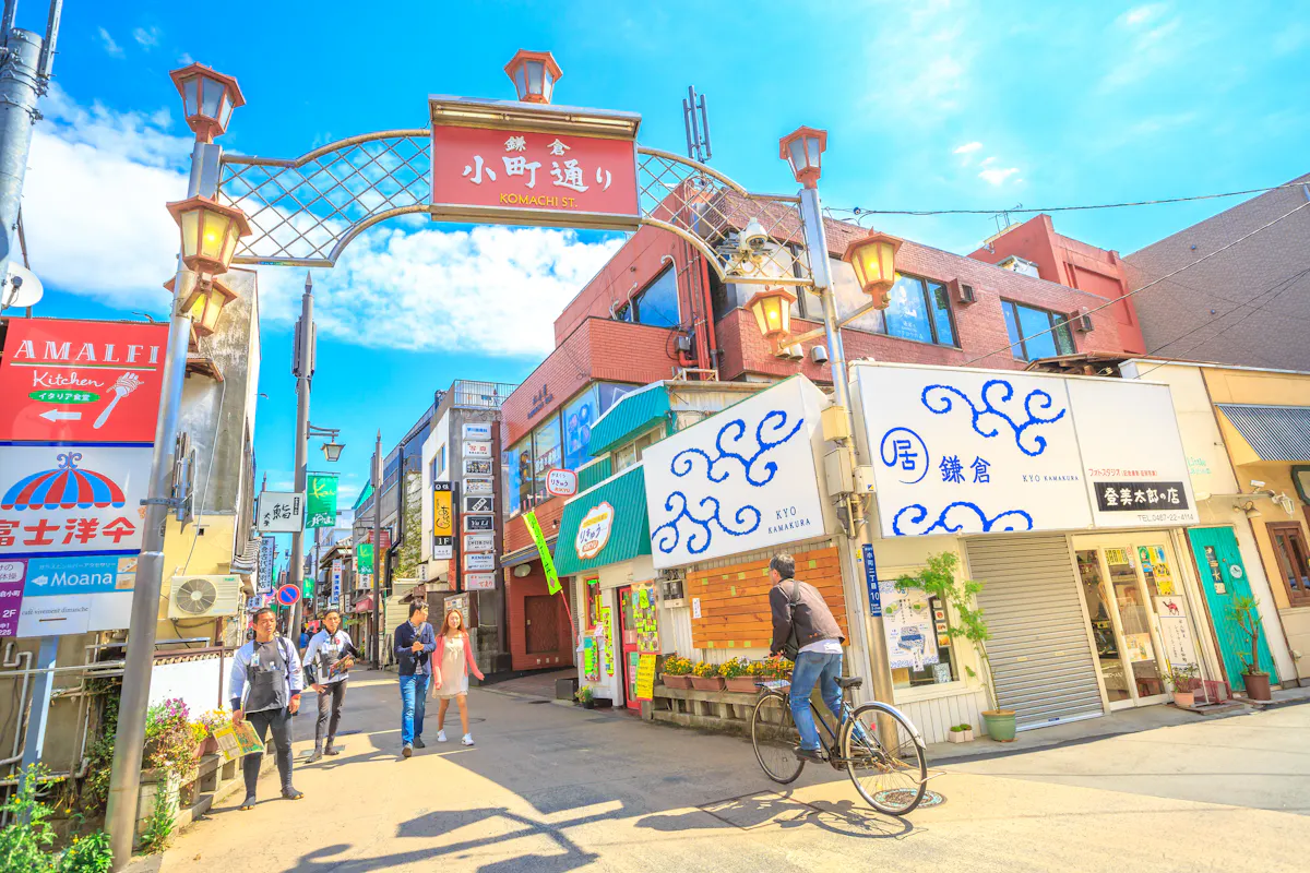 A vibrant street scene in a Japanese town, featuring a cyclist and pedestrians. The area is lined with colorful shops and signs under a decorative archway, with a bright blue sky overhead.