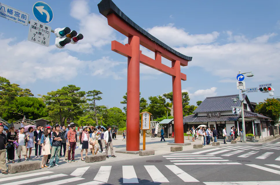 A large red torii gate stands prominently at a crossroads, surrounded by people waiting to cross the street. Behind the gate, a traditional building and lush green trees are visible under a partly cloudy sky. Traffic lights and signs are overhead.