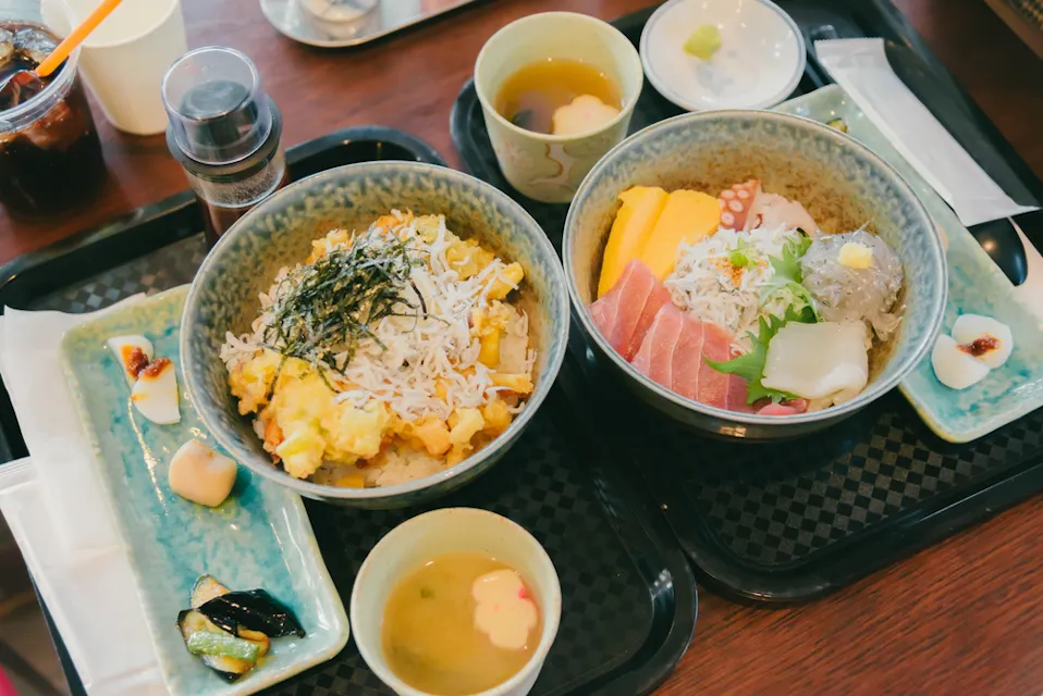 Two Japanese meals on trays, featuring rice bowls topped with various ingredients like seafood, shredded seaweed, and egg. Accompanied by small bowls of miso soup, pickles, a drink, and condiments on a wooden table.