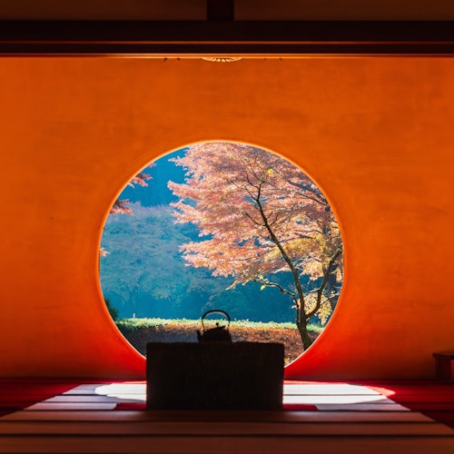 Kamakura in Autumn A view of vibrant autumn leaves and a serene landscape seen through a circular window. The room indoors is dimly lit with traditional decor, featuring a teapot on a platform in the foreground.