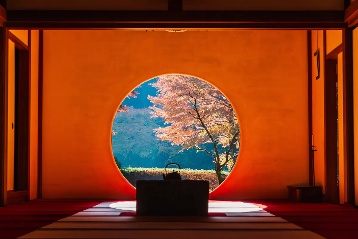 Kamakura in Autumn A view of vibrant autumn leaves and a serene landscape seen through a circular window. The room indoors is dimly lit with traditional decor, featuring a teapot on a platform in the foreground.