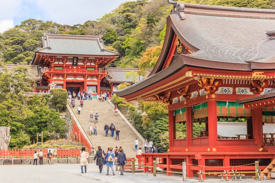 Red and ornate traditional Japanese buildings in a temple complex with people walking along a wide staircase. The background features lush green trees and a clear sky.