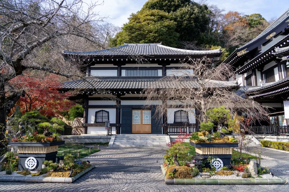 A traditional Japanese building with a tiled roof stands surrounded by trees. In front, there are landscaped gardens with manicured plants and shrubs. Stone pathways lead to the entrance. The scene is peaceful and well-maintained.