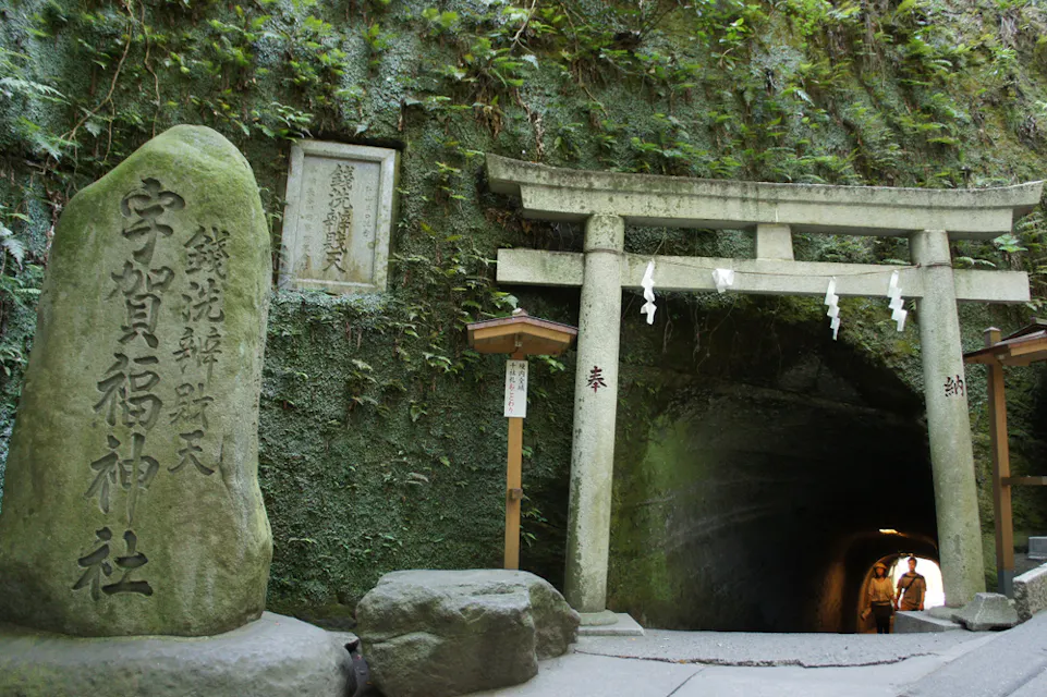 Torii gate at the entrance to a tunnel at Tsurugaoka Hachimangu, Japan. The scene includes a large stone with Japanese inscriptions, a smaller wooden sign, and a few people visible inside the dimly lit tunnel.