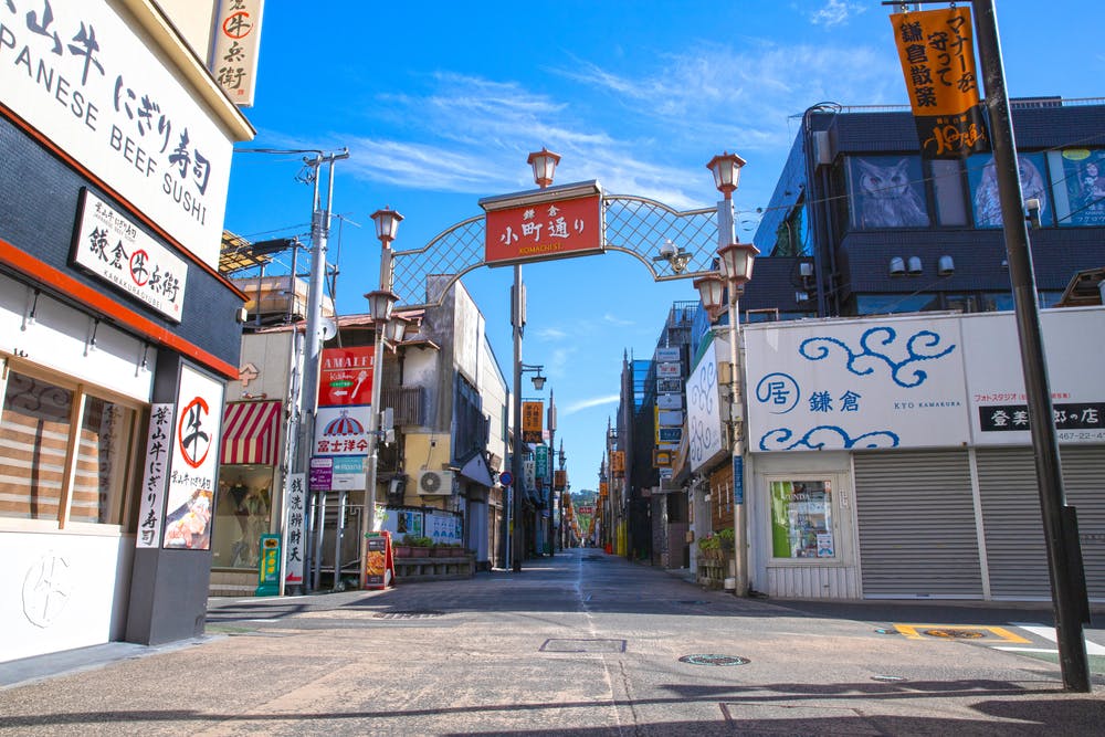 A vibrant, sunny street scene in a Japanese shopping district. A decorative archway spans the walkway, with various shops and signs on both sides. The sky is clear and blue, enhancing the lively atmosphere.