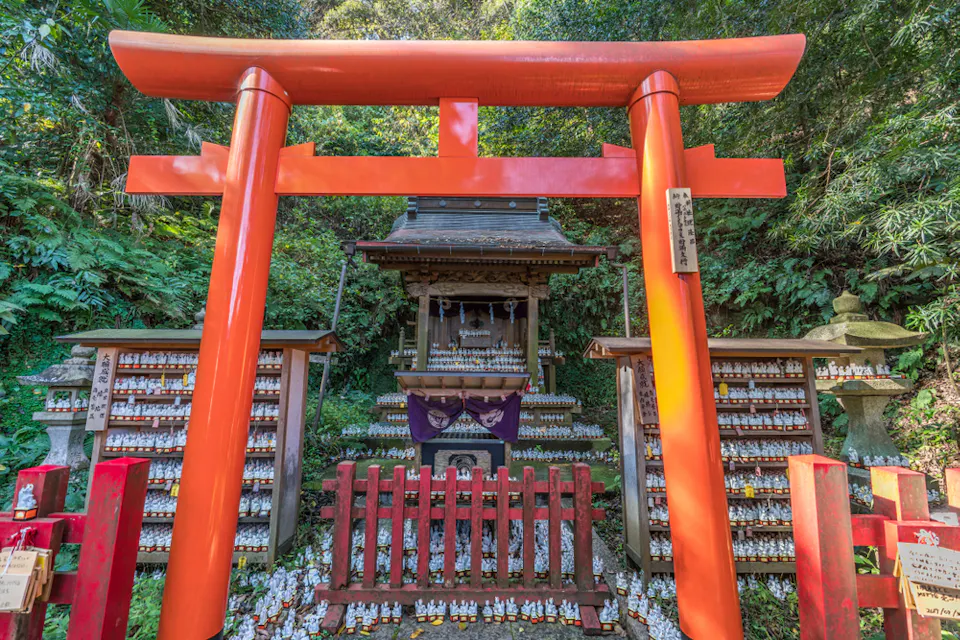 A red torii gate frames a small Shinto shrine surrounded by lush greenery. Miniature fox statues are neatly arranged on shelves to the left and right of the shrine. A stone lantern is partially visible to the side.