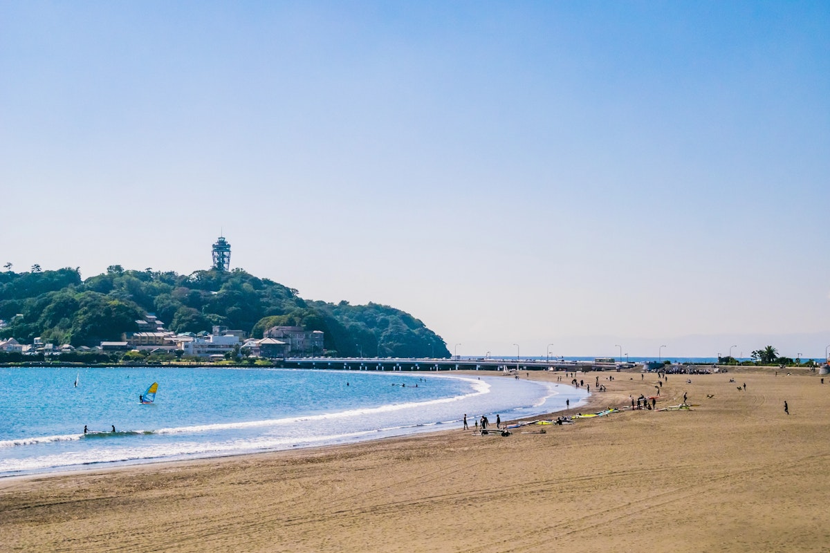 Kamakura Beaches A sandy beach with a few people walking and relaxing. The ocean is calm with windsurfers in the water. In the background, a hill with trees and a tower overlook a bridge extending into the sea under a clear blue sky.