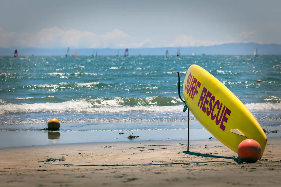 Zaimokuza Beach A bright yellow "Surf Rescue" board stands upright on a sandy beach, with orange buoys nearby. The ocean is calm with gentle waves, and sailboats are visible on the horizon under a clear, sunny sky.