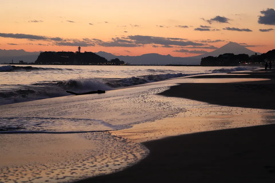 Shichirigahama Beach A serene sunset over a beach with gentle waves lapping against the shore. In the distance, an island with a lighthouse is visible under a colorful sky, and a silhouetted mountain stands on the horizon.