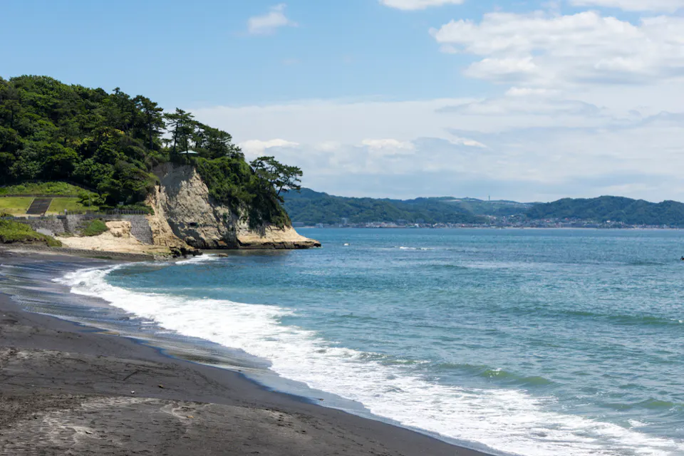 Inamuragasaki Beach Scenic view of a beach with dark sand and gentle waves lapping at the shore. A tree-covered cliff extends into the ocean, with distant hills and a blue sky filled with scattered clouds in the background.