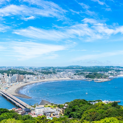 Enoshima Island A scenic view of a coastal city with a curved shoreline, a long bridge, and boats docked nearby. The sky is clear with wispy clouds, and greenery is visible in the foreground, enhancing the vibrant landscape.