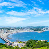 Enoshima Island A scenic view of a coastal city with a curved shoreline, a long bridge, and boats docked nearby. The sky is clear with wispy clouds, and greenery is visible in the foreground, enhancing the vibrant landscape.