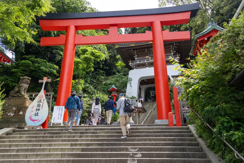 People ascending stone steps toward a red torii gate leading to a traditional Japanese shrine. The scene is surrounded by lush green trees, creating a serene atmosphere. A stone guardian statue and a decorative sign are visible on the left.