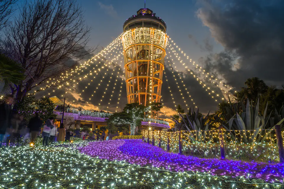 A tall tower illuminated with strings of golden lights is surrounded by a garden decorated with vibrant purple and white lights. The sky is dusky with scattered clouds, creating a festive and enchanting evening scene.