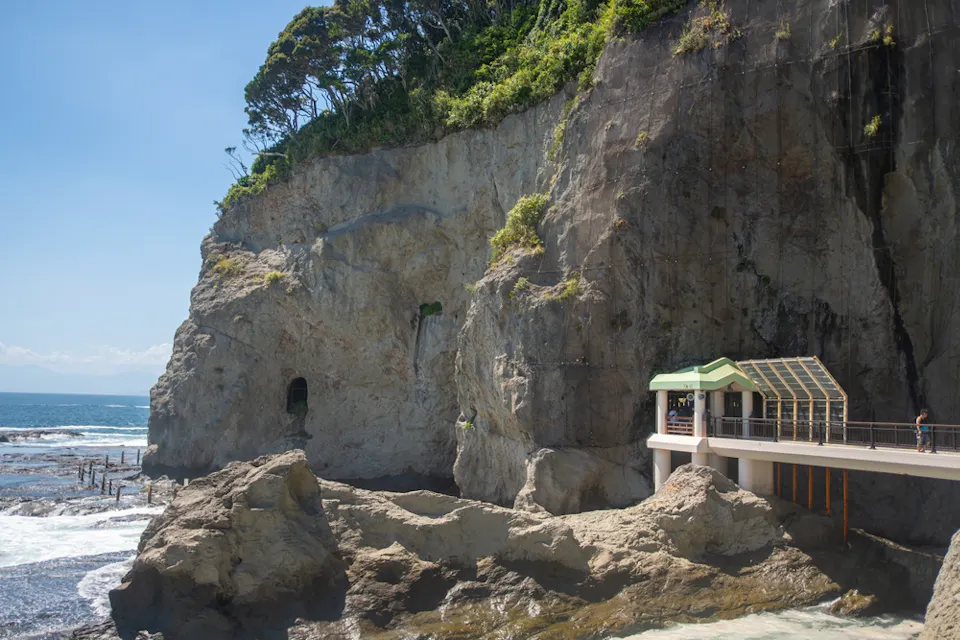 A scenic view of a rocky coastal area featuring a pathway with a shelter. Lush greenery tops the cliffs, and the ocean waves crash against the rocks. The sky is clear and sunny, enhancing the tranquil atmosphere.