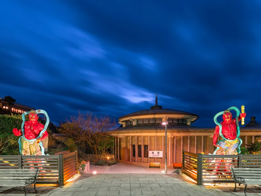 Two vibrant red statues stand at the entrance of a circular building with a traditional design. The statues are positioned on either side of a gated walkway. The evening sky above is deep blue, with clouds creating a dynamic backdrop.