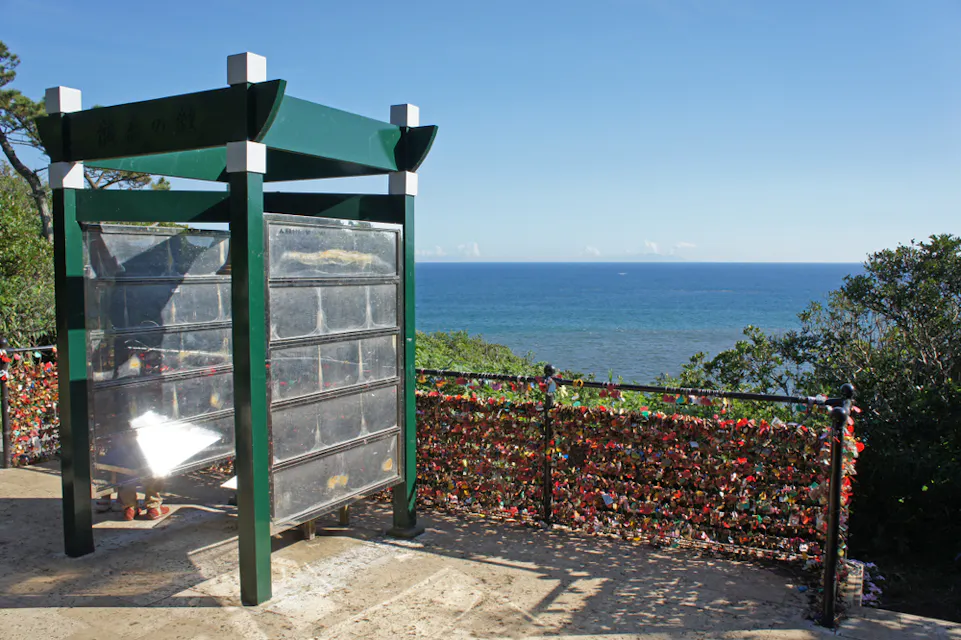 A scenic viewpoint overlooking a vast blue ocean, framed by lush greenery. A structure with a green roof stands on the left, and a fence adorned with colorful locks stretches across the scene. The sky is clear and bright.