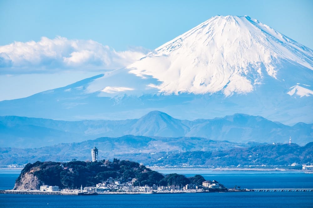 A scenic view of Mount Fuji with a snow-capped peak under a clear blue sky. In the foreground, an island with a lighthouse and small buildings sits in a calm, blue body of water.