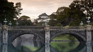 A historic stone bridge with ornate lamps spans a calm moat, with a traditional Japanese building and lush trees in the background at sunset.