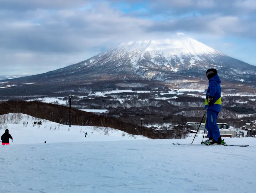 A skier in a blue outfit stands on a snow-covered slope, looking at a distant snow-capped mountain under a partly cloudy sky. Other skiers can be seen in the background.