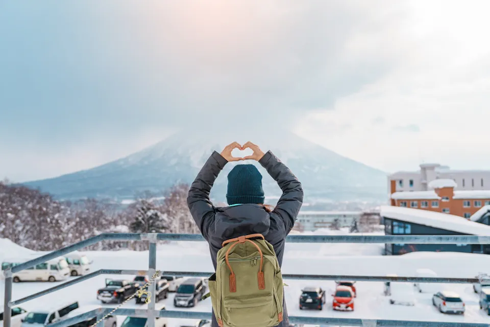 A person in a winter coat and hat stands on a snowy rooftop, forming a heart shape with their hands. They face a snow-covered mountain under a cloudy sky, with parked cars and buildings visible in the background.