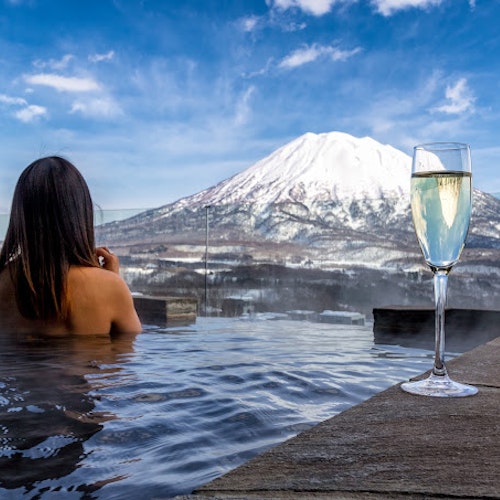The Vale Niseko A person relaxes in an outdoor hot spring pool, overlooking a snow-covered mountain. A glass of champagne sits on the edge of the pool. The sky is clear with a few clouds.