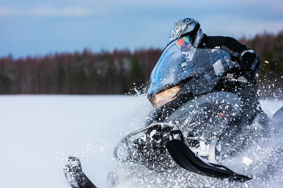 A person riding a snowmobile at high speed on a snowy landscape. Snow is flying up around the vehicle, and a line of trees is visible in the background against a blue sky. The rider is wearing a helmet and protective gear.