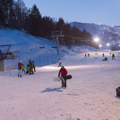 Night view of Niseko ski resort at Niseko People enjoy skiing and snowboarding on a gently lit ski slope in the early evening. Snow-covered trees and mountains surround the area. A group is preparing near the ski lift while others are gliding down the hill.