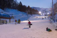 People enjoy skiing and snowboarding on a gently lit ski slope in the early evening. Snow-covered trees and mountains surround the area. A group is preparing near the ski lift while others are gliding down the hill.