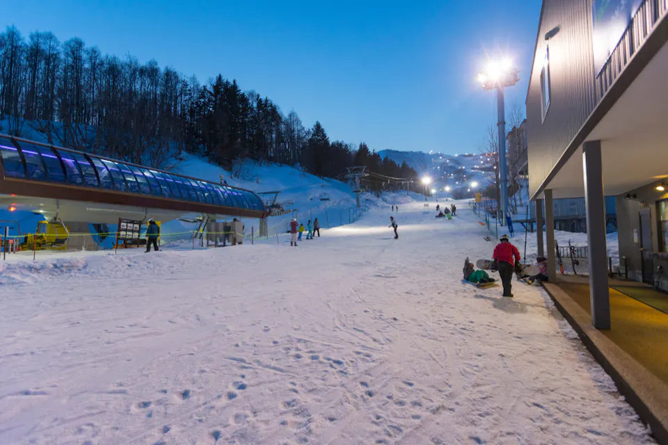 A snowy ski resort at dusk with people preparing to ski. A ski lift is on the left, and a building is on the right. The area is illuminated by bright lights, with trees and hills in the background under a clear blue sky.