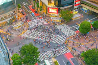 Aerial view of a bustling city intersection with a large crowd of pedestrians crossing multiple zebra stripes. Surrounding buildings have bright advertisements, and greenery adds contrast to the urban setting.