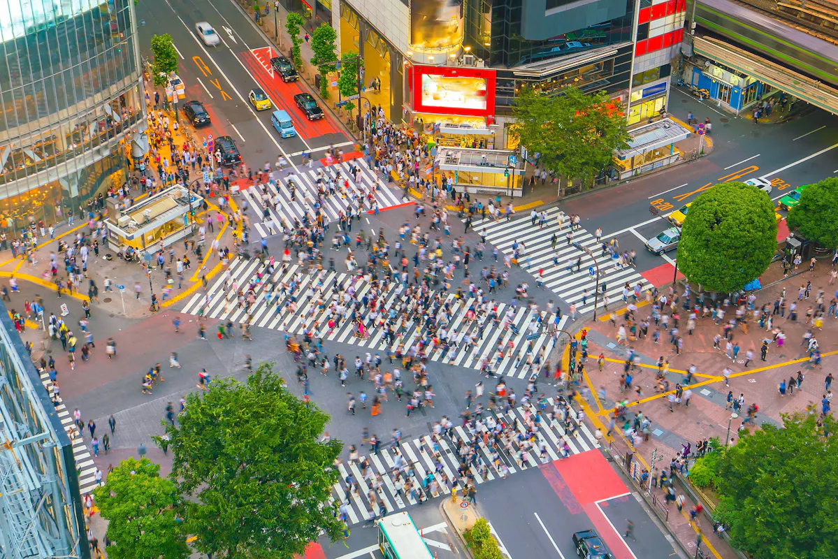 Aerial view of a bustling city intersection with a large crowd of pedestrians crossing multiple zebra stripes. Surrounding buildings have bright advertisements, and greenery adds contrast to the urban setting.