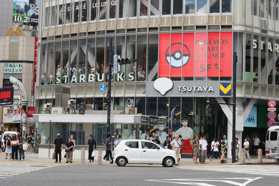 Busy city street scene with people crossing in front of a Starbucks and Tsutaya store. A white car is waiting at the intersection. Large digital ads and signs, including one for Pokémon, are visible on a multi-story building.