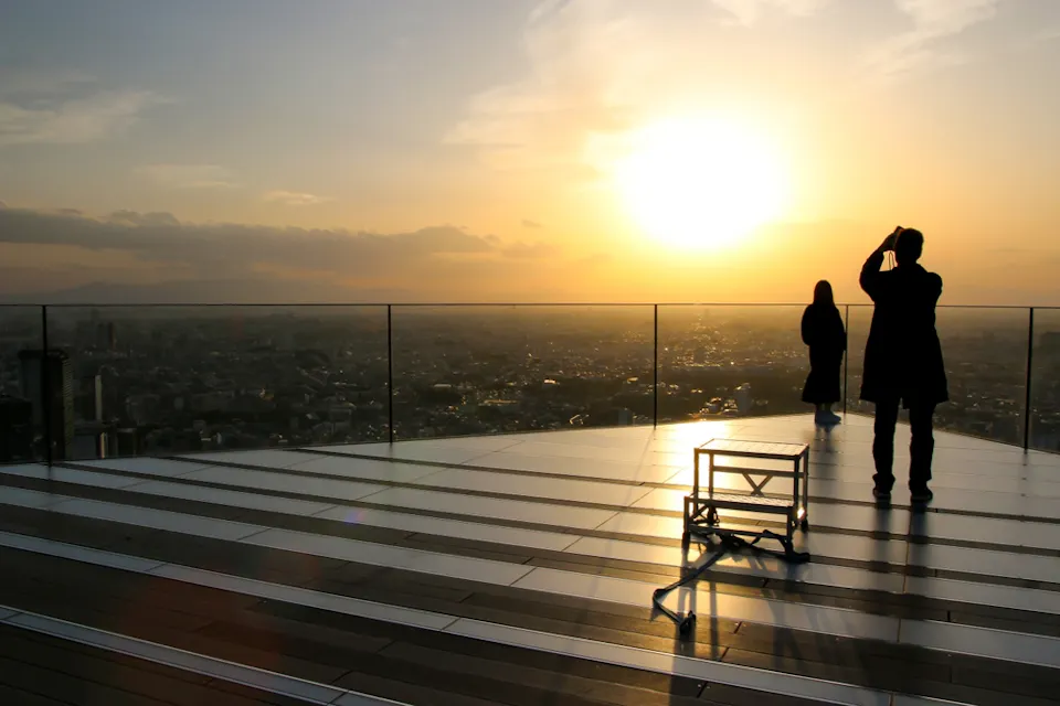 Two people stand on a rooftop deck at sunset, silhouetted against the bright sky. One person is taking a photo with a smartphone, while the other gazes into the distance. The cityscape stretches out below, partially obscured by haze.