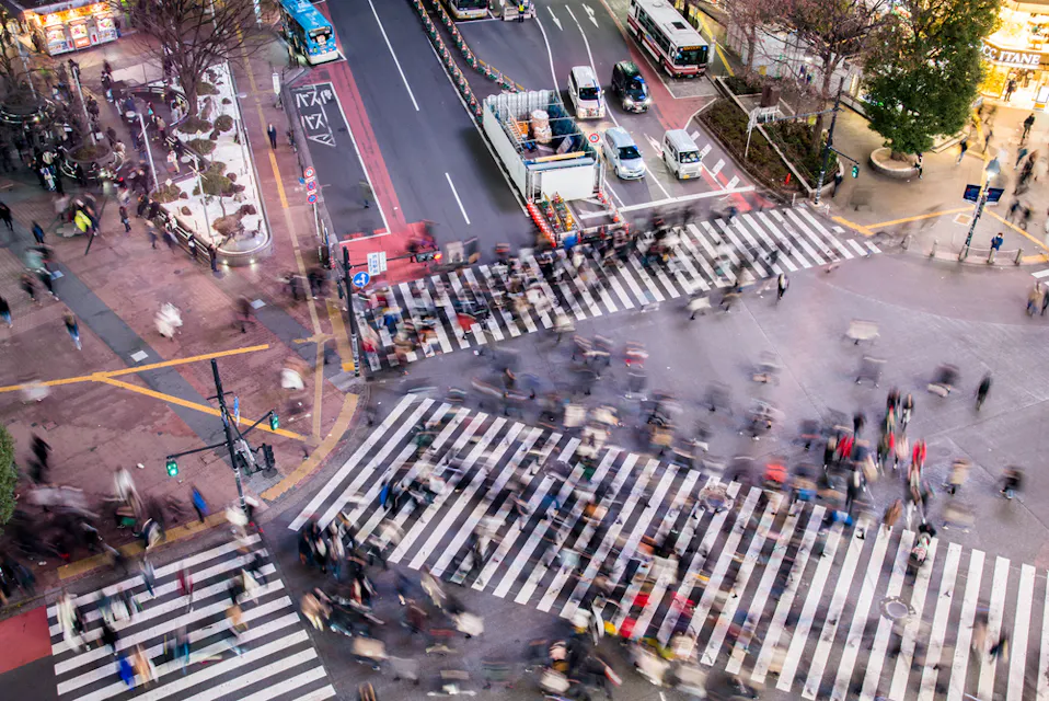 Aerial view of a busy city intersection with blurred motion of pedestrians crossing multiple striped crosswalks. Streets are lined with parked vehicles and surrounded by trees and buildings. The image conveys a sense of bustling urban life.