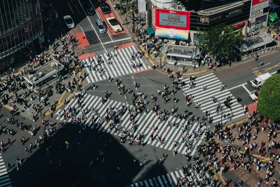 Aerial view of a busy city intersection with numerous pedestrians crossing multiple crosswalks. Buildings and signs surround the area, and shadows from nearby structures cast across the scene.
