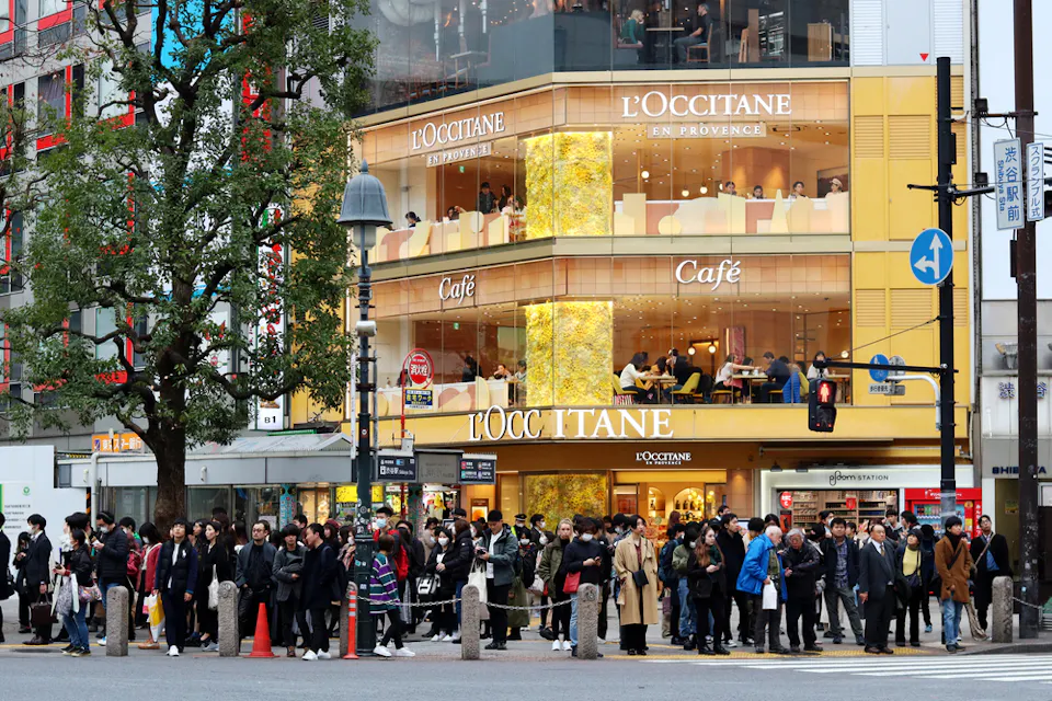 A large crowd of people waits at a busy city crosswalk in front of a multi-story L'Occitane store with prominent signage. The building has glass windows with people visible inside the café on the second floor.