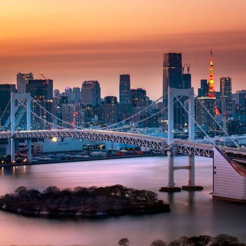 Odaiba A cityscape at dusk featuring a prominent suspension bridge over the water. The skyline behind includes tall buildings with one tower illuminated in red and orange hues. The sky is a gradient of orange and purple, reflecting on the water's surface.