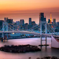Odaiba A cityscape at dusk featuring a prominent suspension bridge over the water. The skyline behind includes tall buildings with one tower illuminated in red and orange hues. The sky is a gradient of orange and purple, reflecting on the water's surface.