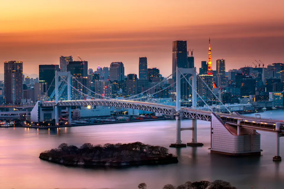 A cityscape at dusk featuring a prominent suspension bridge over the water. The skyline behind includes tall buildings with one tower illuminated in red and orange hues. The sky is a gradient of orange and purple, reflecting on the water's surface.